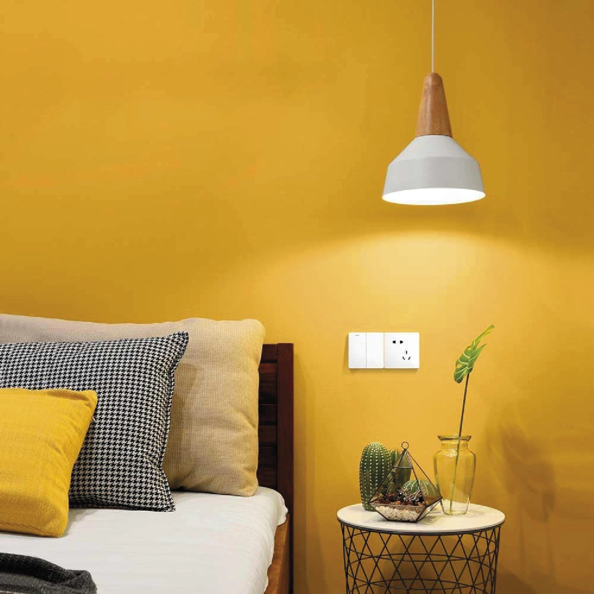 Yellow wall with a white pendant light, bed with pillows, and small table with plants.