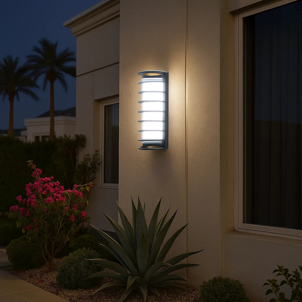 Outdoor wall light fixture on a building exterior with plants and palm trees in the background.