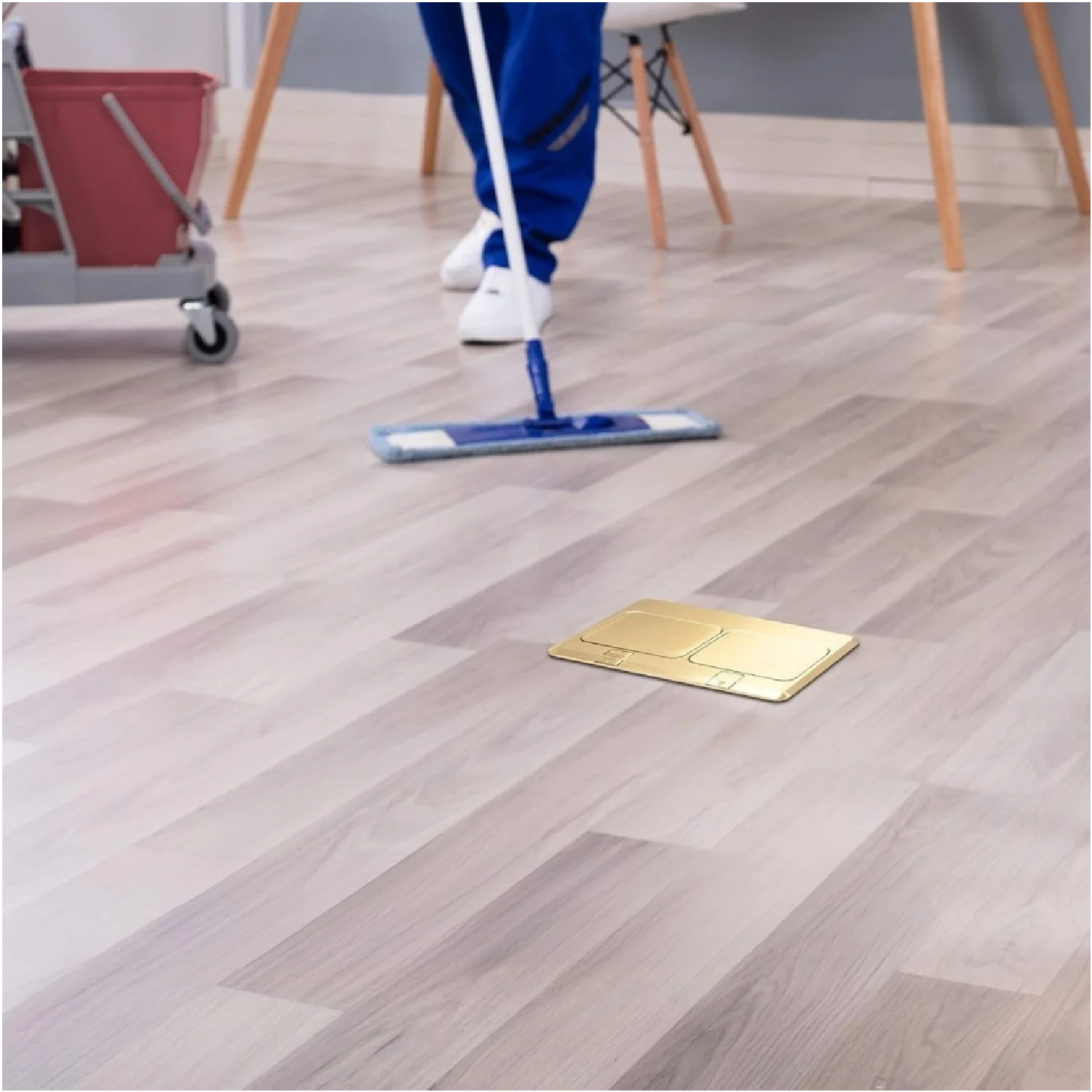 Person cleaning a wooden floor with a mop, with a gold-colored square object on the floor.