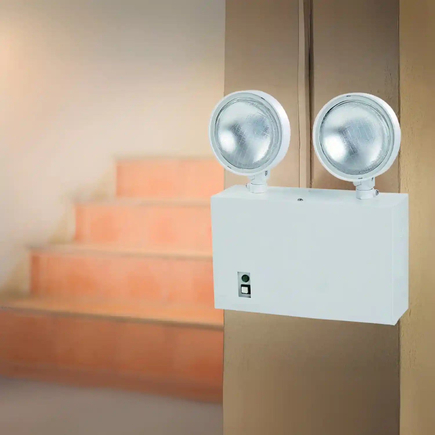 Double-lamp emergency light fixture mounted on a wall with stairs in the background.