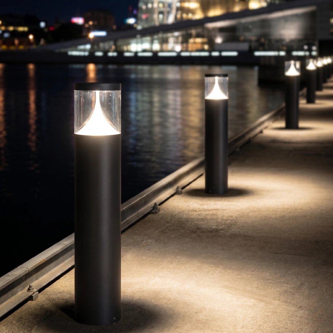 Row of illuminated cylindrical lights along a waterfront pathway at night.