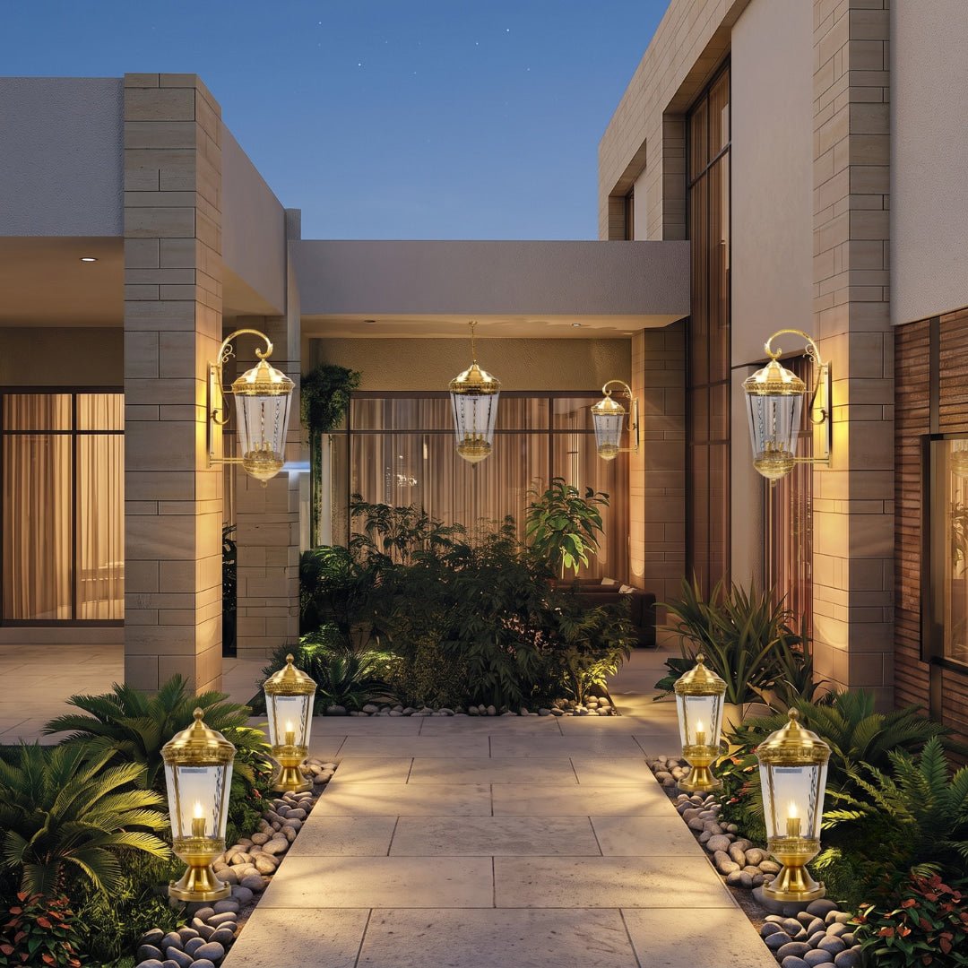 Outdoor pathway lighting set in a modern courtyard at dusk, featuring multiple gold and clear glass lantern-style wall and path lights illuminating a stone walkway and landscaped greenery.