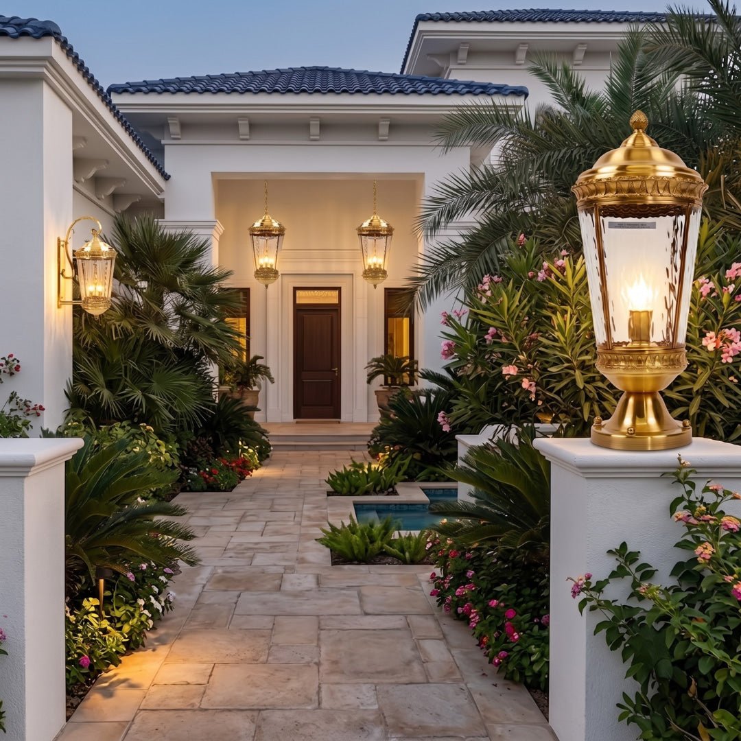 A beautifully lit outdoor pathway leading to a white stucco house entrance at dusk, featuring a prominent ornate gold and glass lantern on a white pillar in the foreground, with palm trees, pink flowers, and warm ambient lighting.