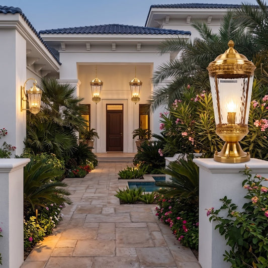 A beautifully lit outdoor pathway leading to a white stucco house entrance at dusk, featuring a prominent ornate gold and glass lantern on a white pillar in the foreground, with palm trees, pink flowers, and warm ambient lighting.