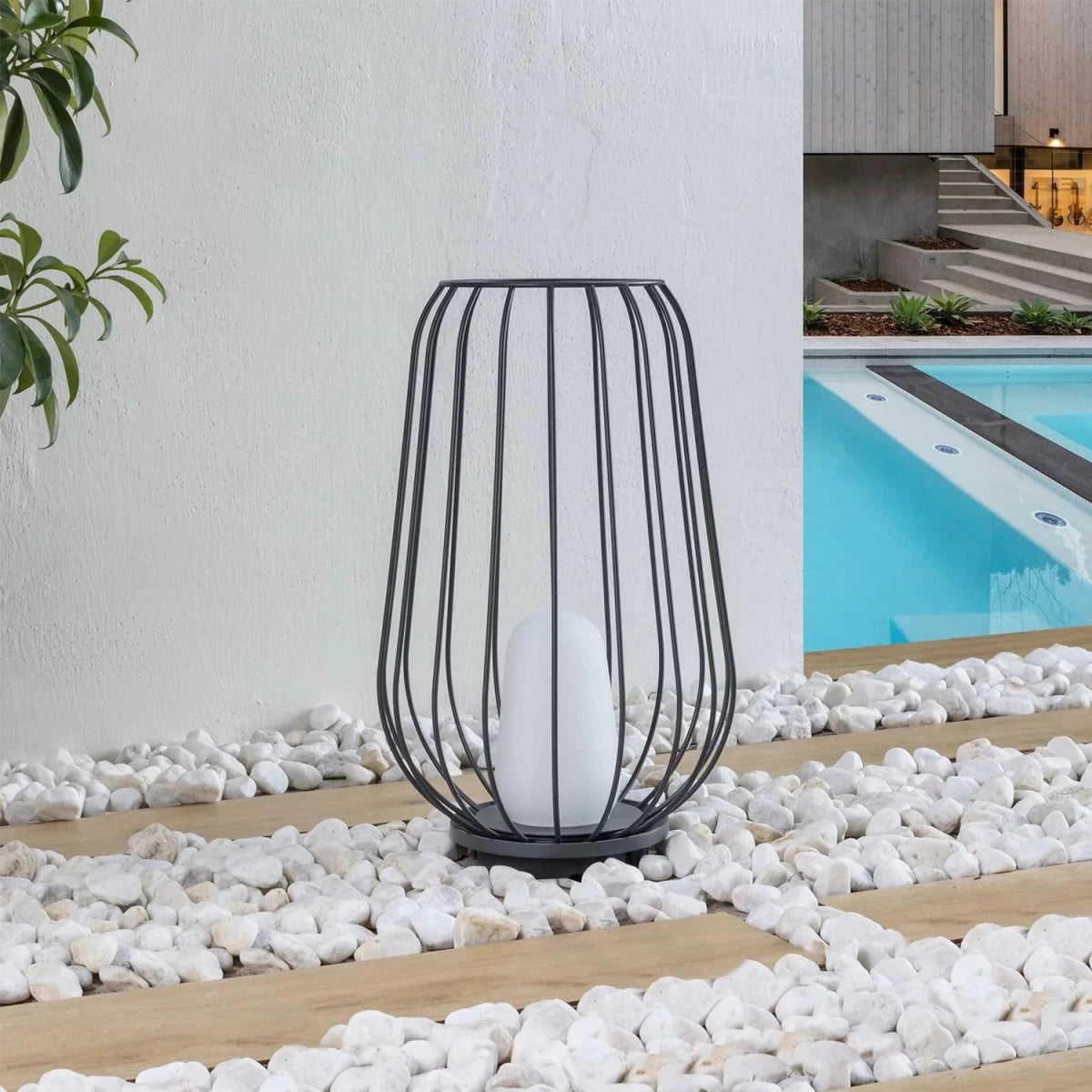 Black wire lantern with a white candle on pebbles near a pool