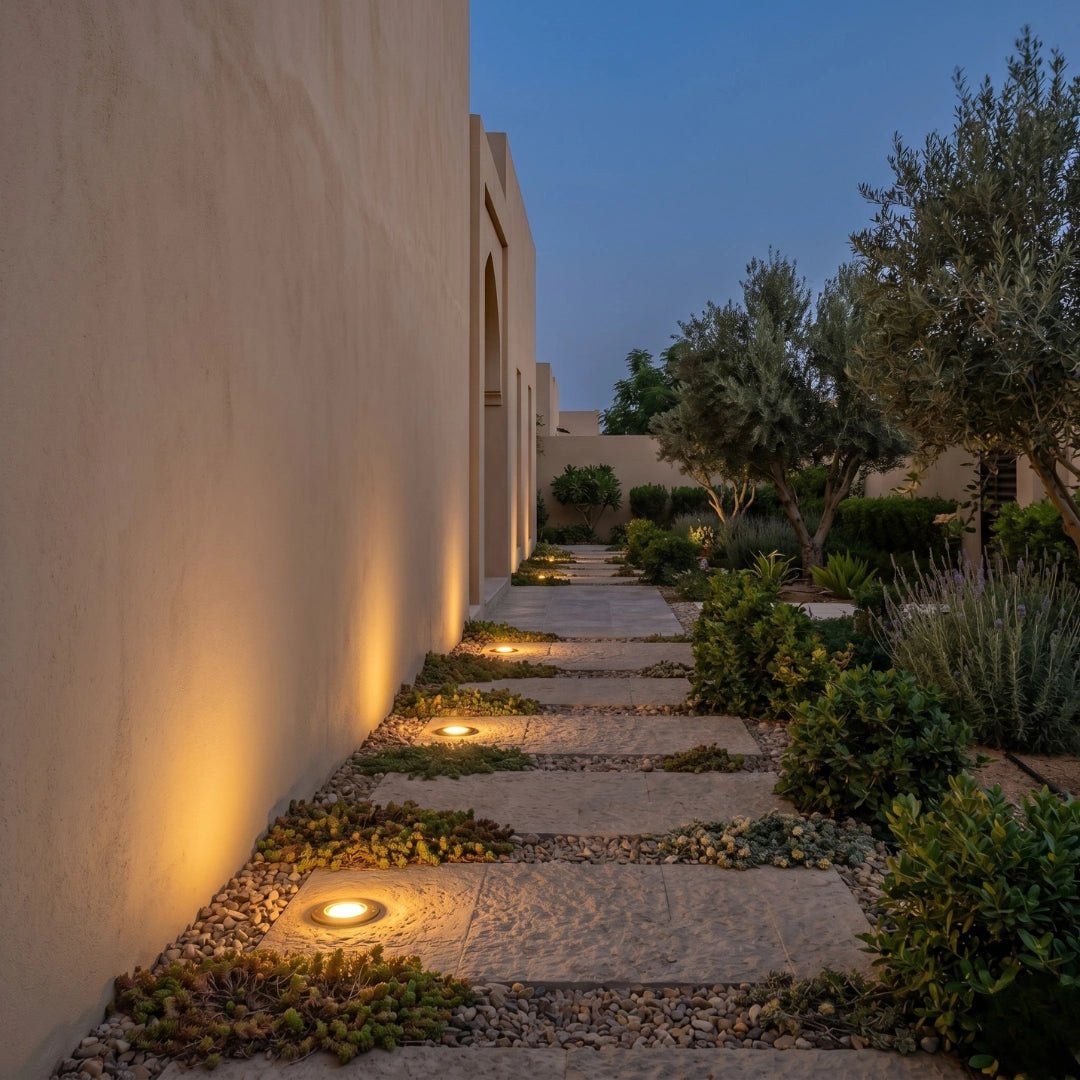 Pathway illuminated by embedded lights next to a building with trees and plants in the background.