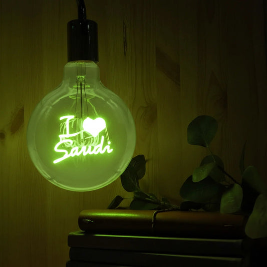 Glowing light bulb with 'I love Saudi' text on a wooden surface with a plant.