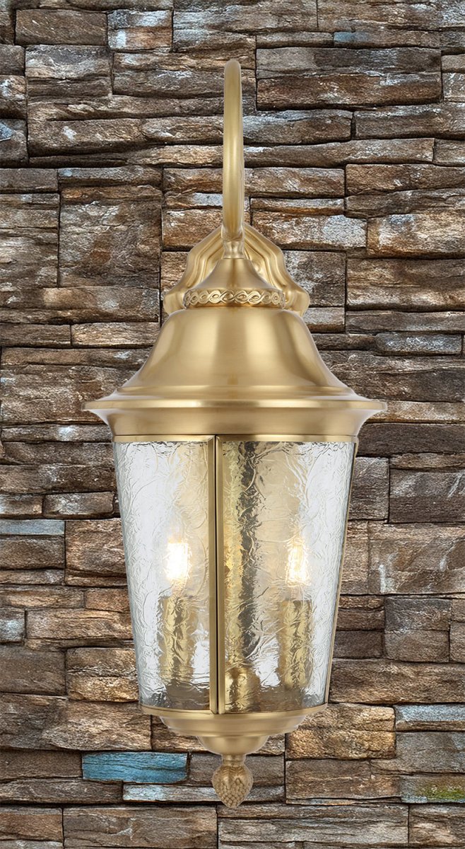 Decorative outdoor light fixture with gold finish and clear glass against a stone wall.