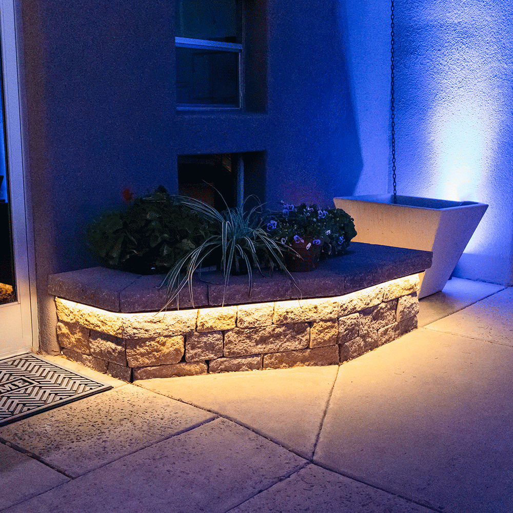 Outdoor seating area with stone bench, plants, and illuminated wall at night.